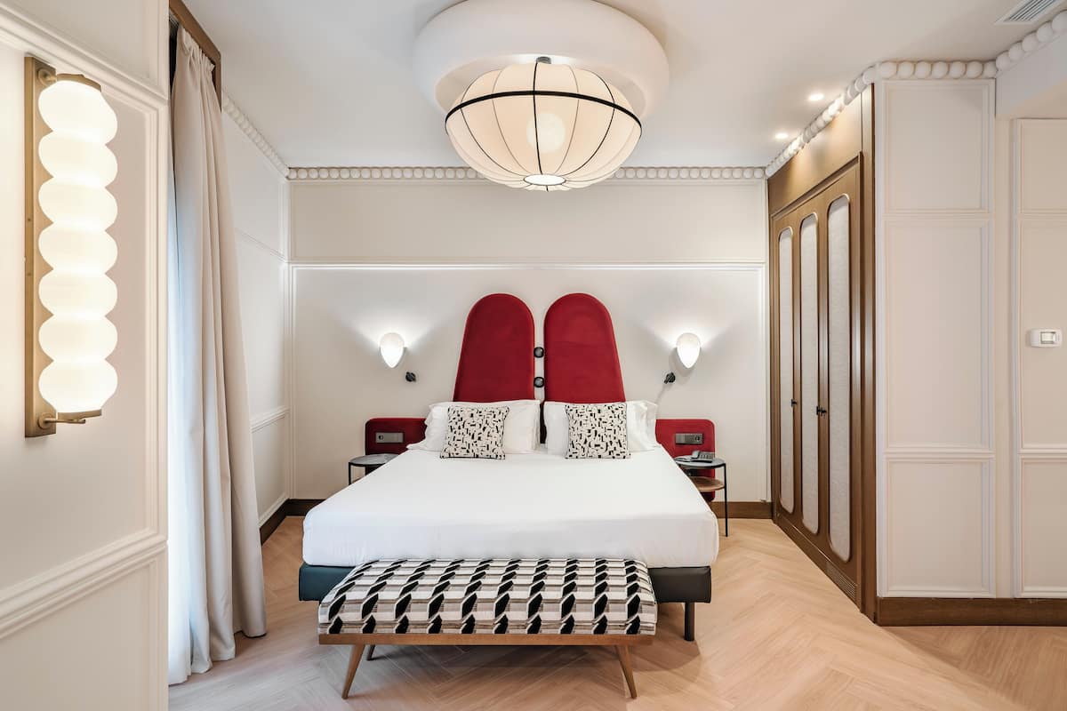 Une chambre d’hôtel avec un grand lit, un banc au pied du lit, un lustre et une armoire.