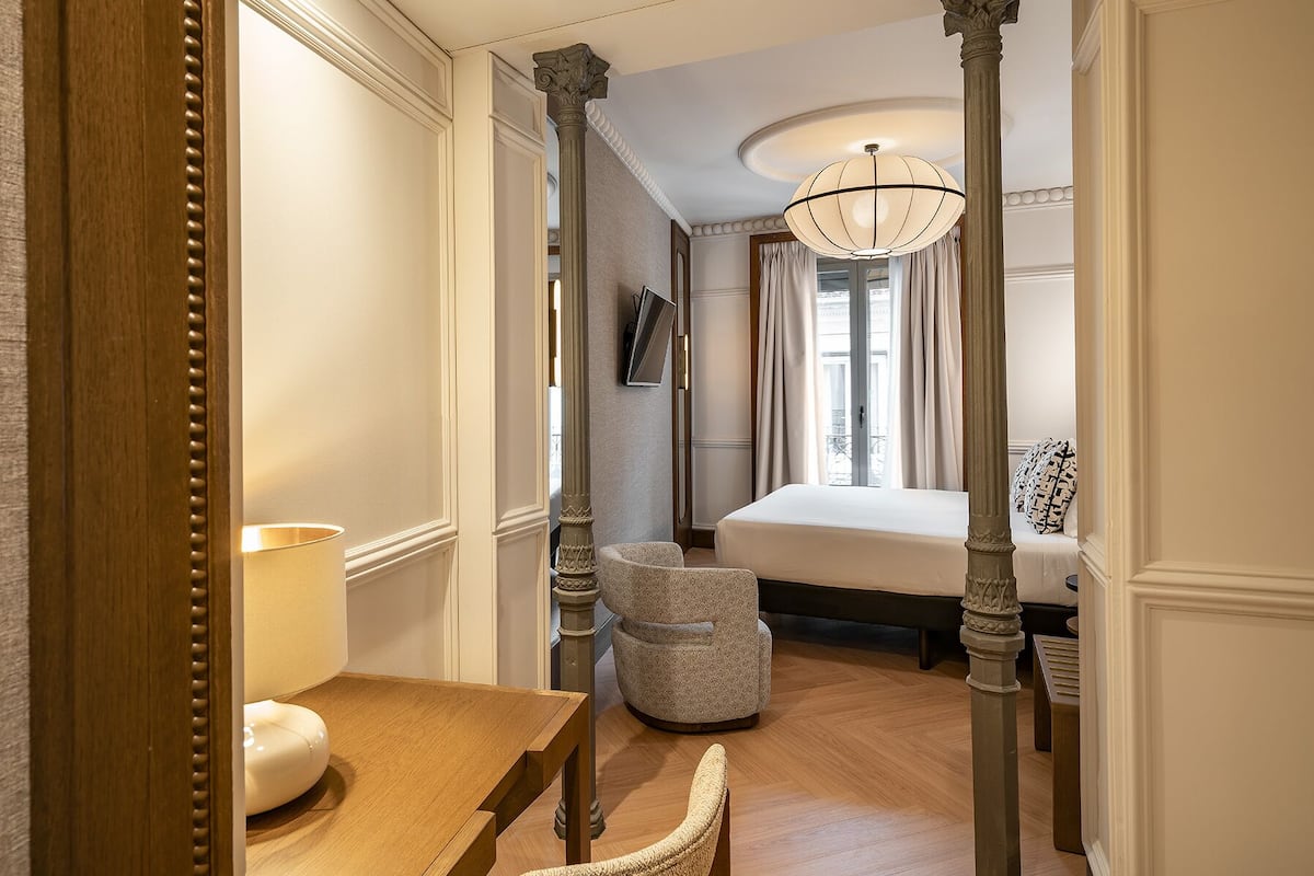 Une chambre d’hôtel avec un bureau en bois, une chaise, un lit, une grande fenêtre avec des rideaux et un lustre.
