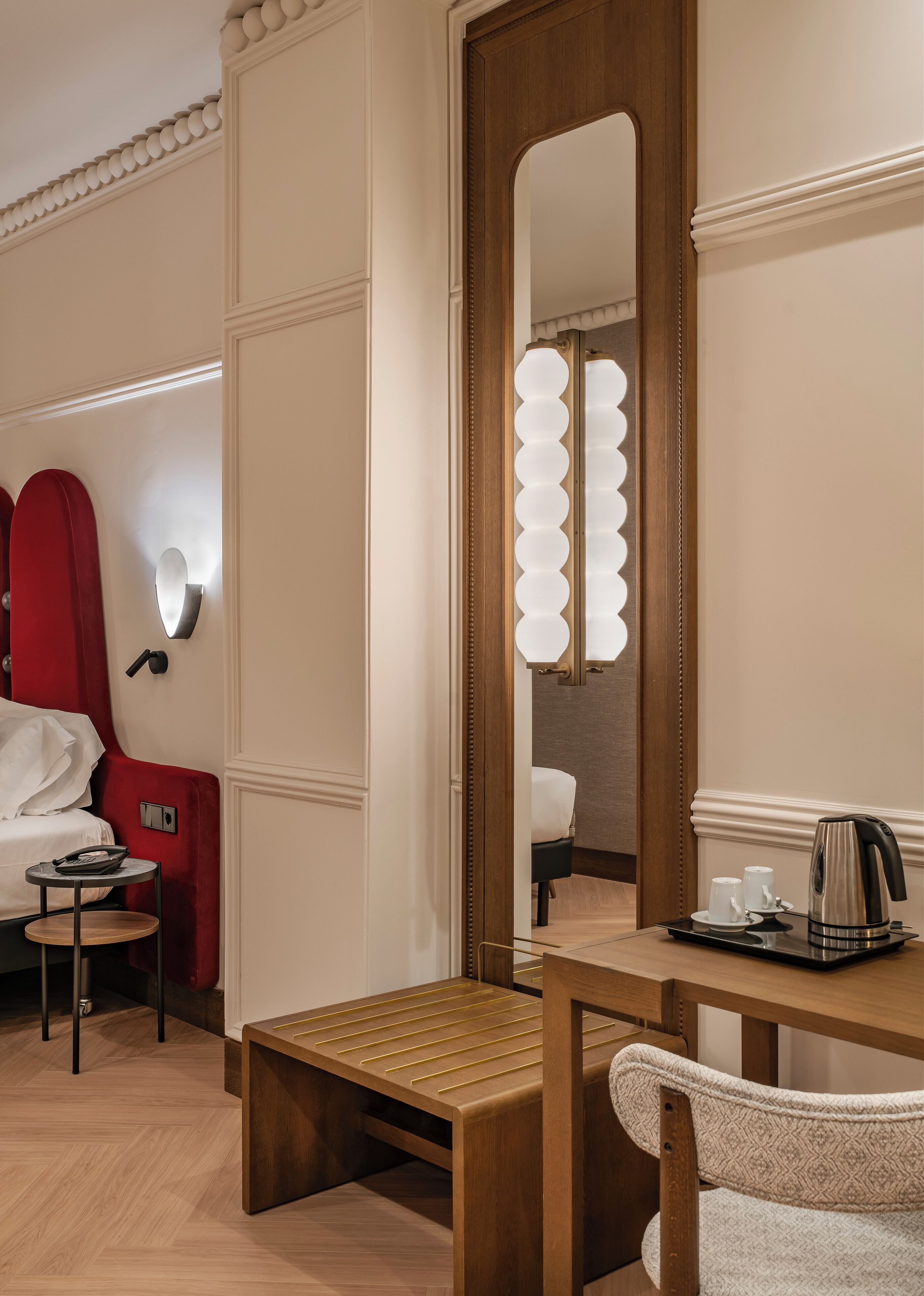 A hotel room with a wooden vanity, a red chair, a small table with a telephone, and a tray with a kettle and cups.