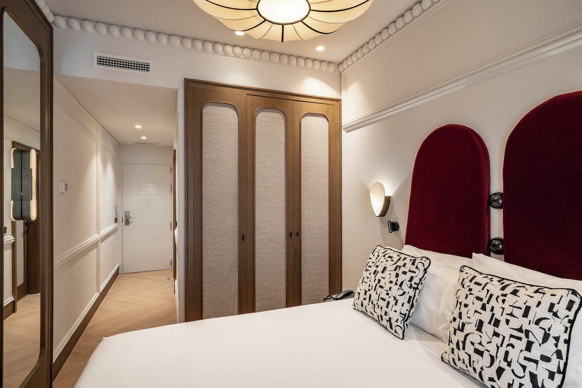 Une chambre d’hôtel avec un grand lit, une armoire, un miroir et une tête de lit rembourrée de tissu rouge.