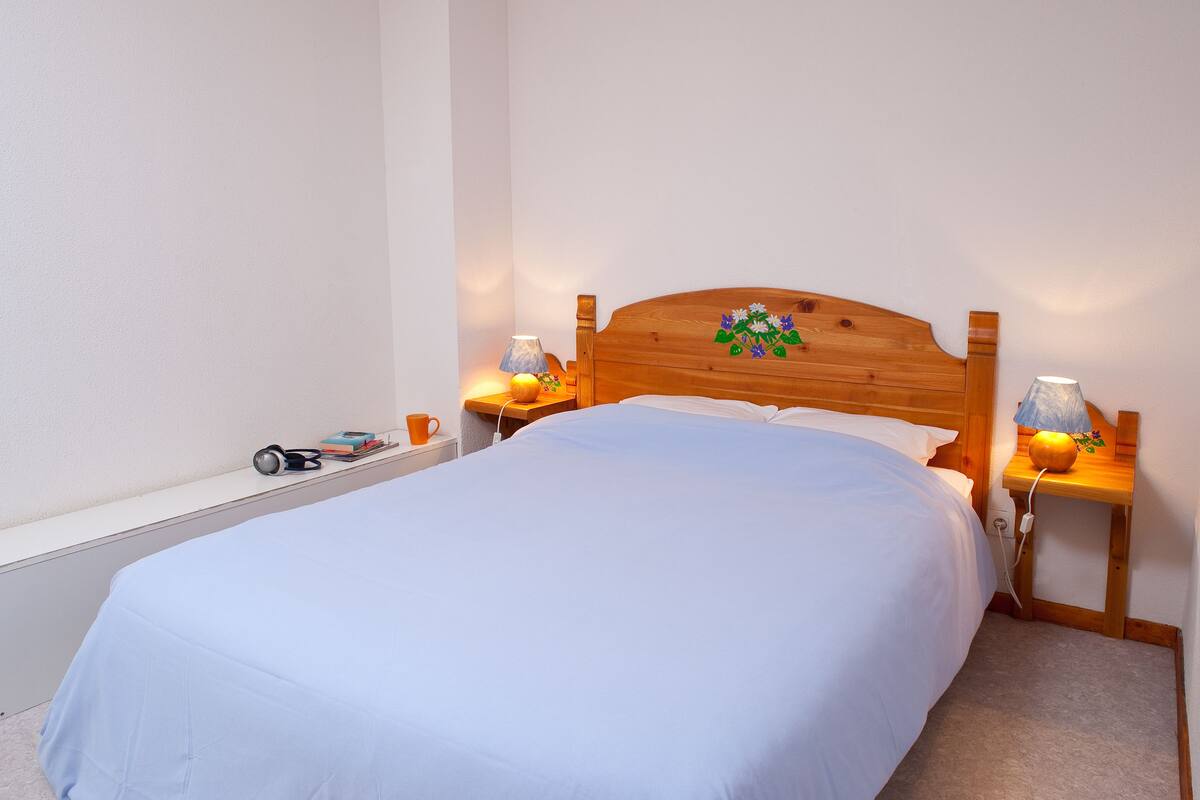 A neatly made bed with a blue bedspread, two bedside lamps, a nightstand with a lamp, a small table with a pumpkin, and a toy car on the floor.