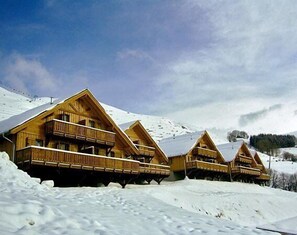 A row of wooden chalets with balconies, set against a snowy mountain backdrop. - Résidence et Chalets de la Fontaine du roi  (Saint-Jean-d'Arves)