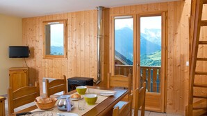 A wooden-paneled room with a dining table set for two, a fireplace, and a view of mountains through large windows. - Résidence et Chalets de la Fontaine du roi  (Saint-Jean-d'Arves)