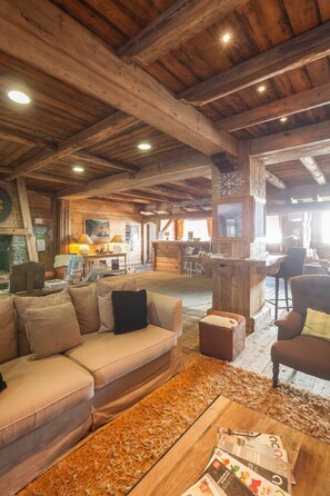 L’intérieur d’un chalet rustique, avec un plafond en bois, un canapé, une table basse avec des magazines et un coin repas comprenant une table et des chaises.