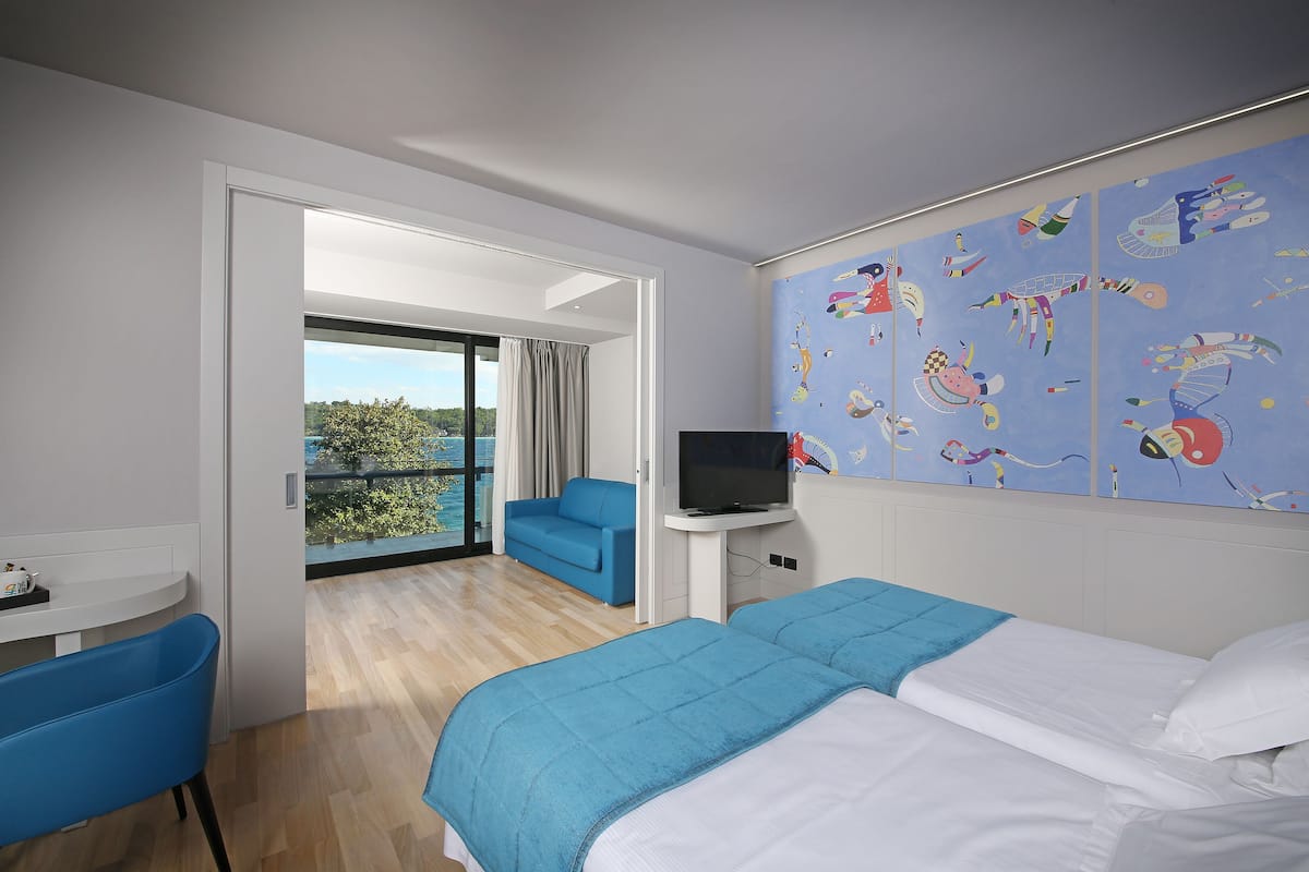 A hotel room with a bed, a TV, a blue chair, a balcony with a view, and a colorful mural on the wall.