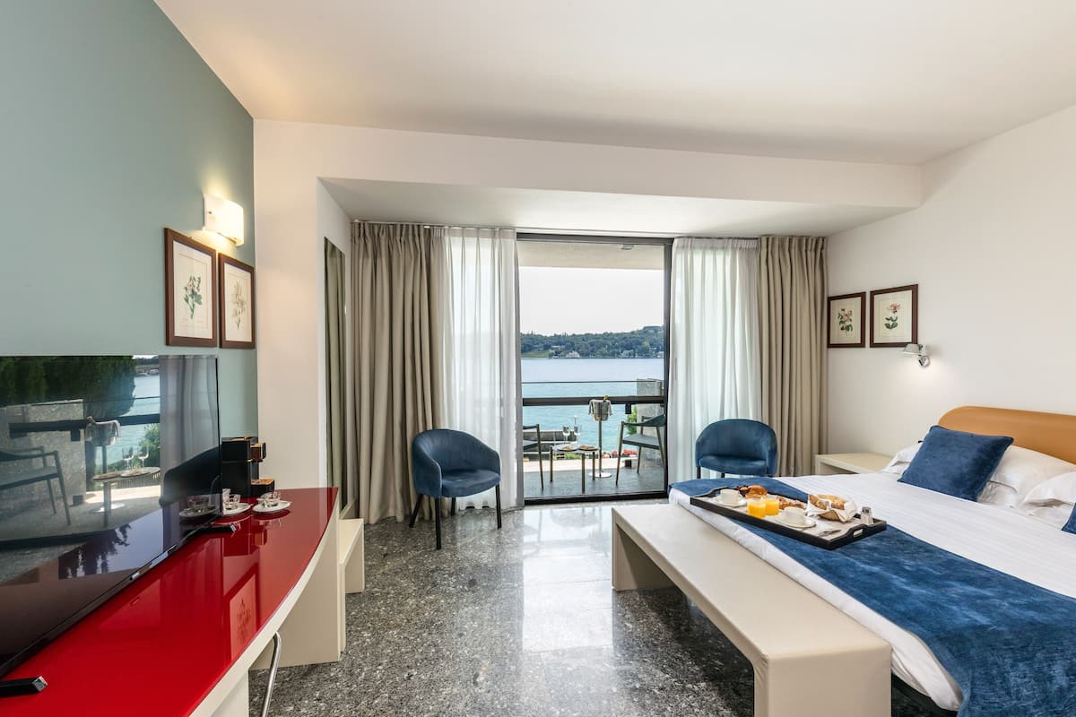 A hotel room with a bed, a desk with a TV, a balcony with a table and chairs, and a view of the sea.