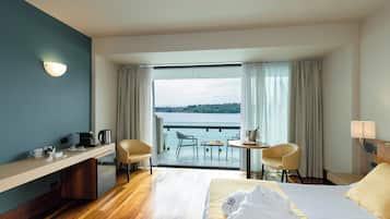 Une chambre d’hôtel comprenant un lit, un bureau, une chaise, une petite table avec une lampe, et donnant sur un plan d’eau grâce à une grande fenêtre.