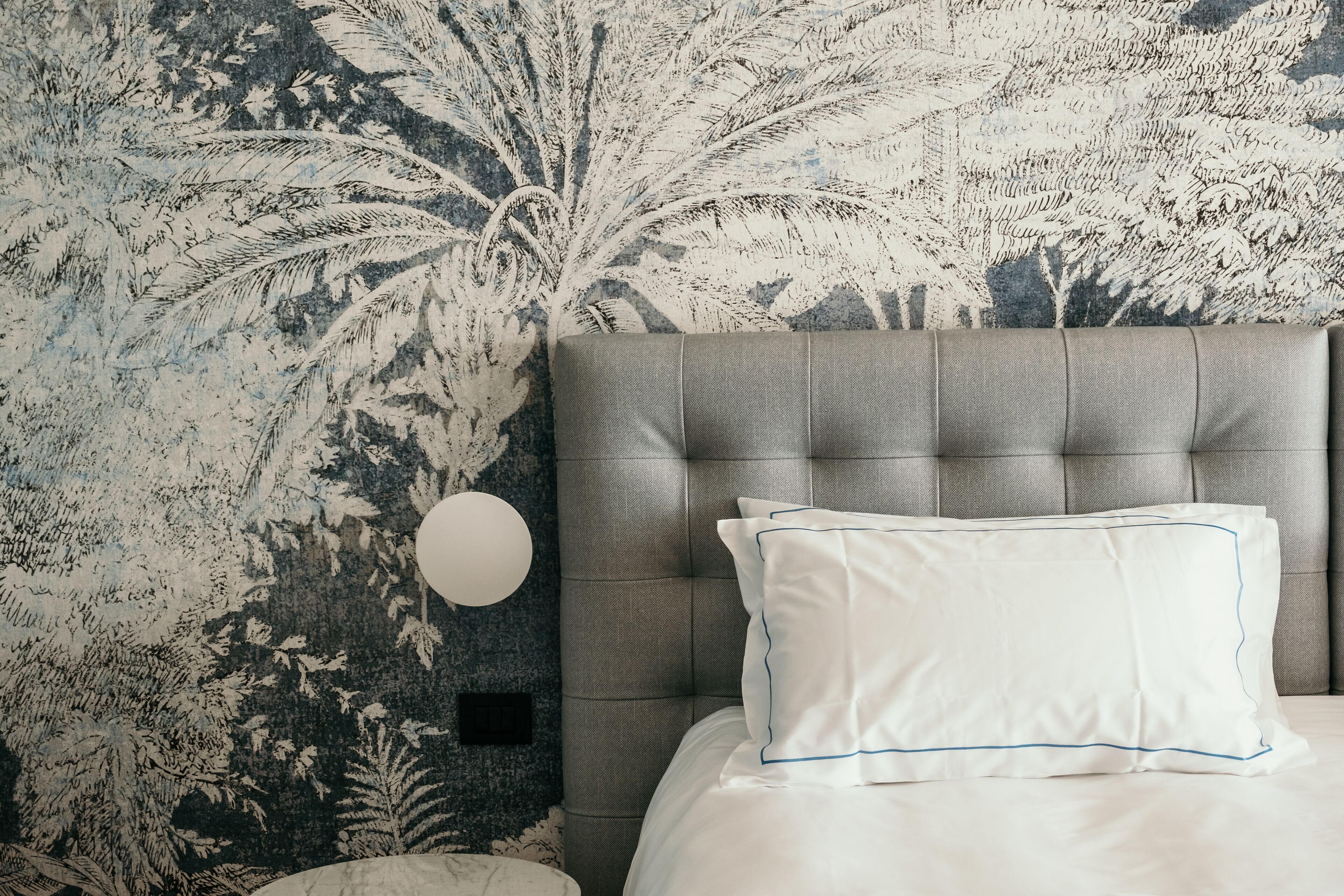 A bedroom with a tufted headboard, a round wall light, and a bed with white bedding and blue-lined pillows.
