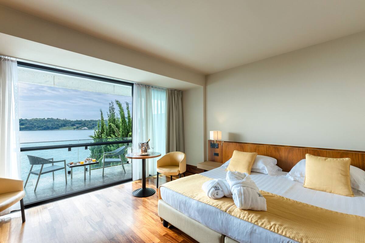 A hotel room with a large window, a bed with white linens, a wooden headboard, and a small table with a lamp.
