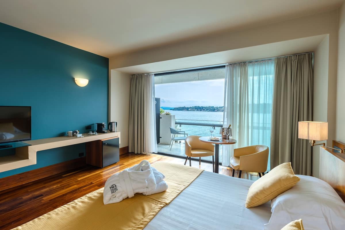 A hotel room with a bed, a desk, a chair, a TV, and a view of the sea.