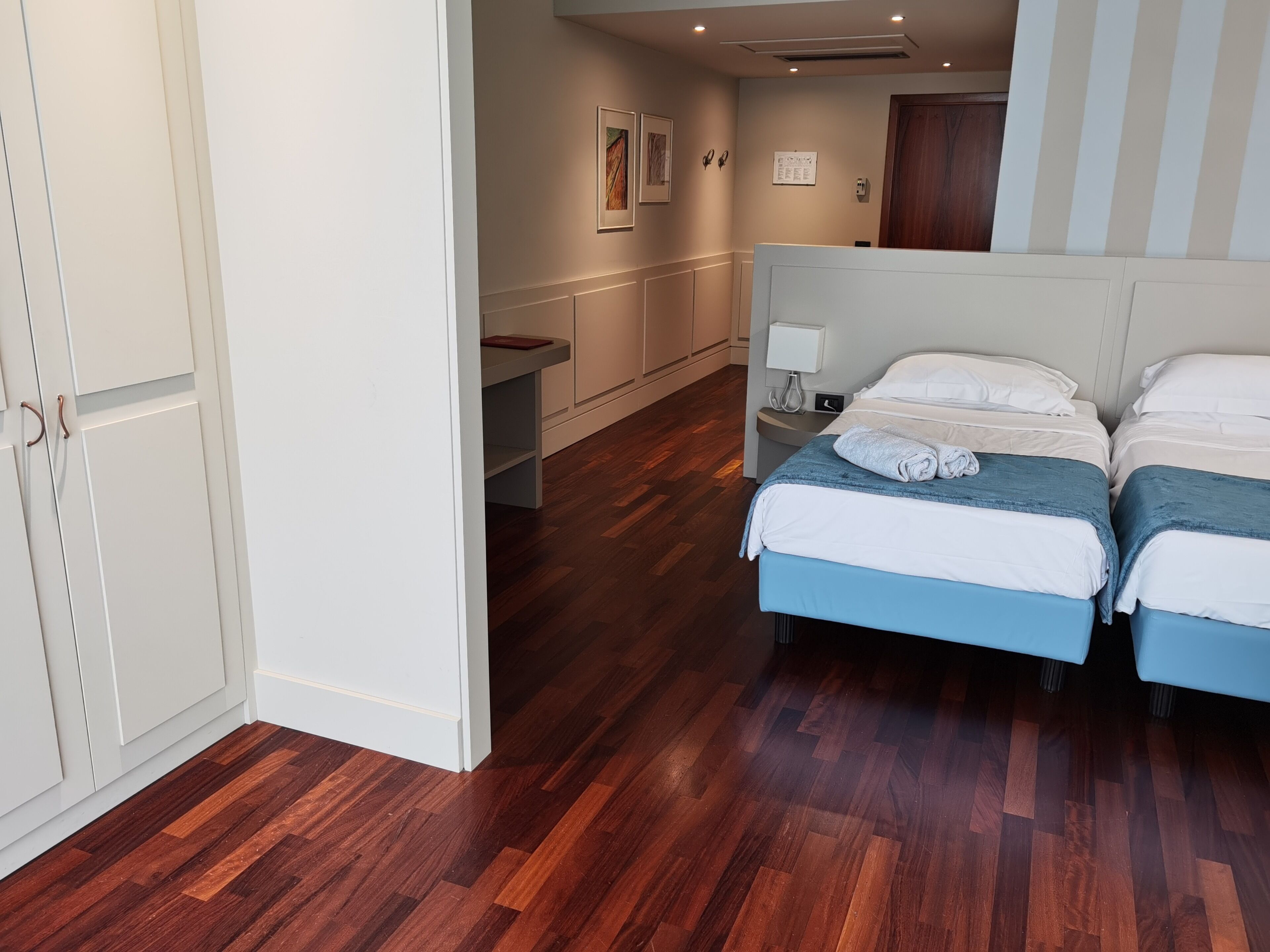 Quarto de hotel com duas camas, uma escrivaninha, uma cadeira, um criado-mudo com abajur e um guarda-roupa.
