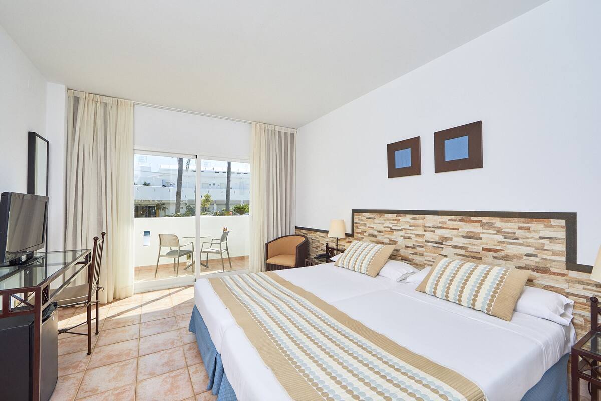 A hotel room with a large bed, a TV, a desk with a chair, and a balcony with a chair.