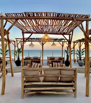 Una pérgola de madera con una lámpara colgante, asientos acolchados y plantas en macetas, con vista a la playa.