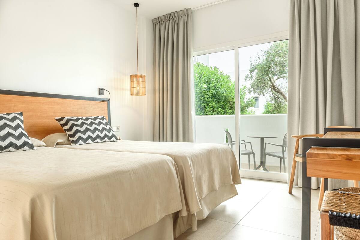 A hotel room with a large bed, two pillows, a bedside table, a lamp, and a view of the outdoors through a sliding glass door.