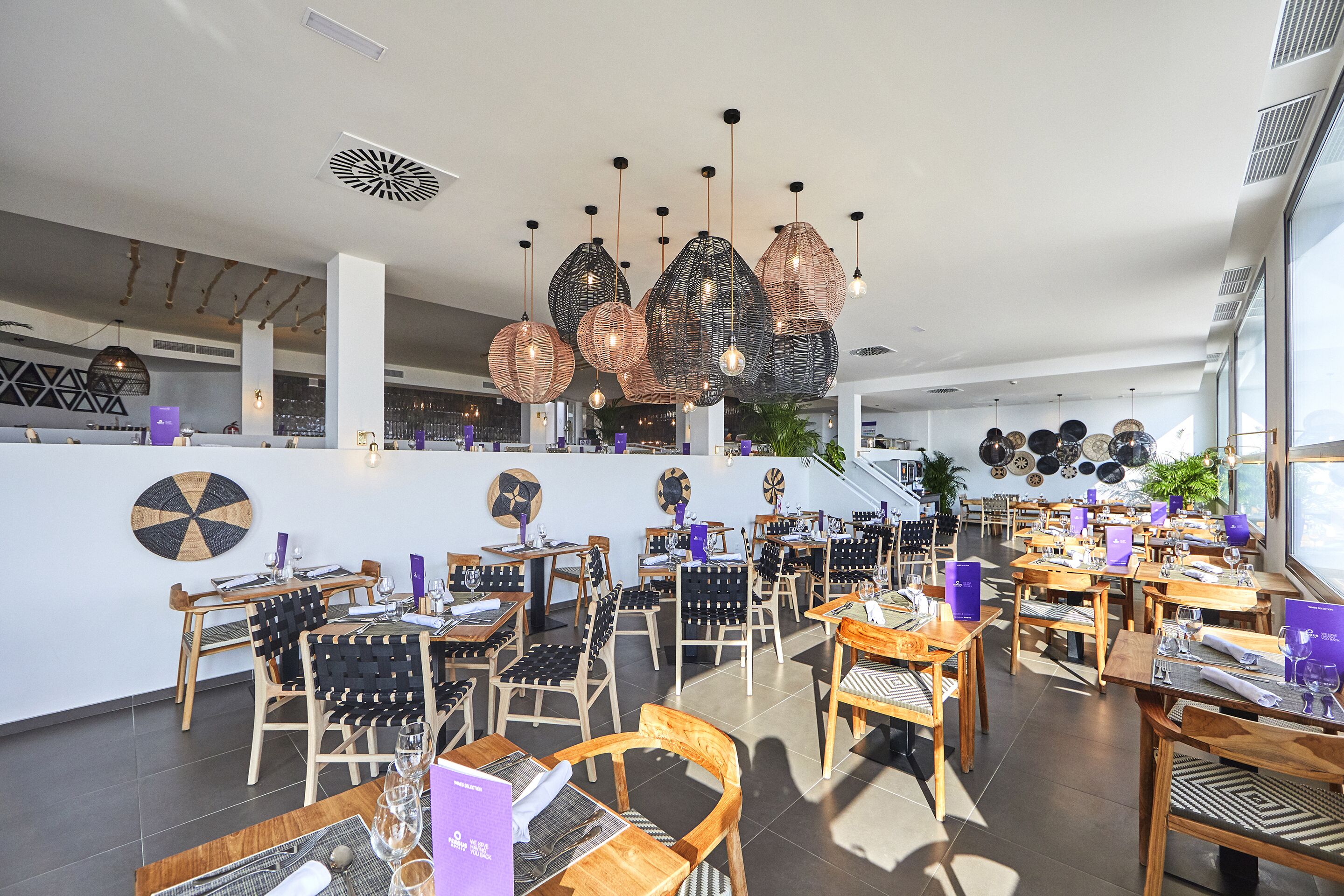 A spacious restaurant with wooden tables and chairs, hanging rattan chandeliers, and a modern interior design.