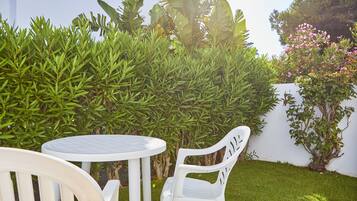 A white outdoor seating area with a round table and two chairs, surrounded by lush greenery and a white wall.