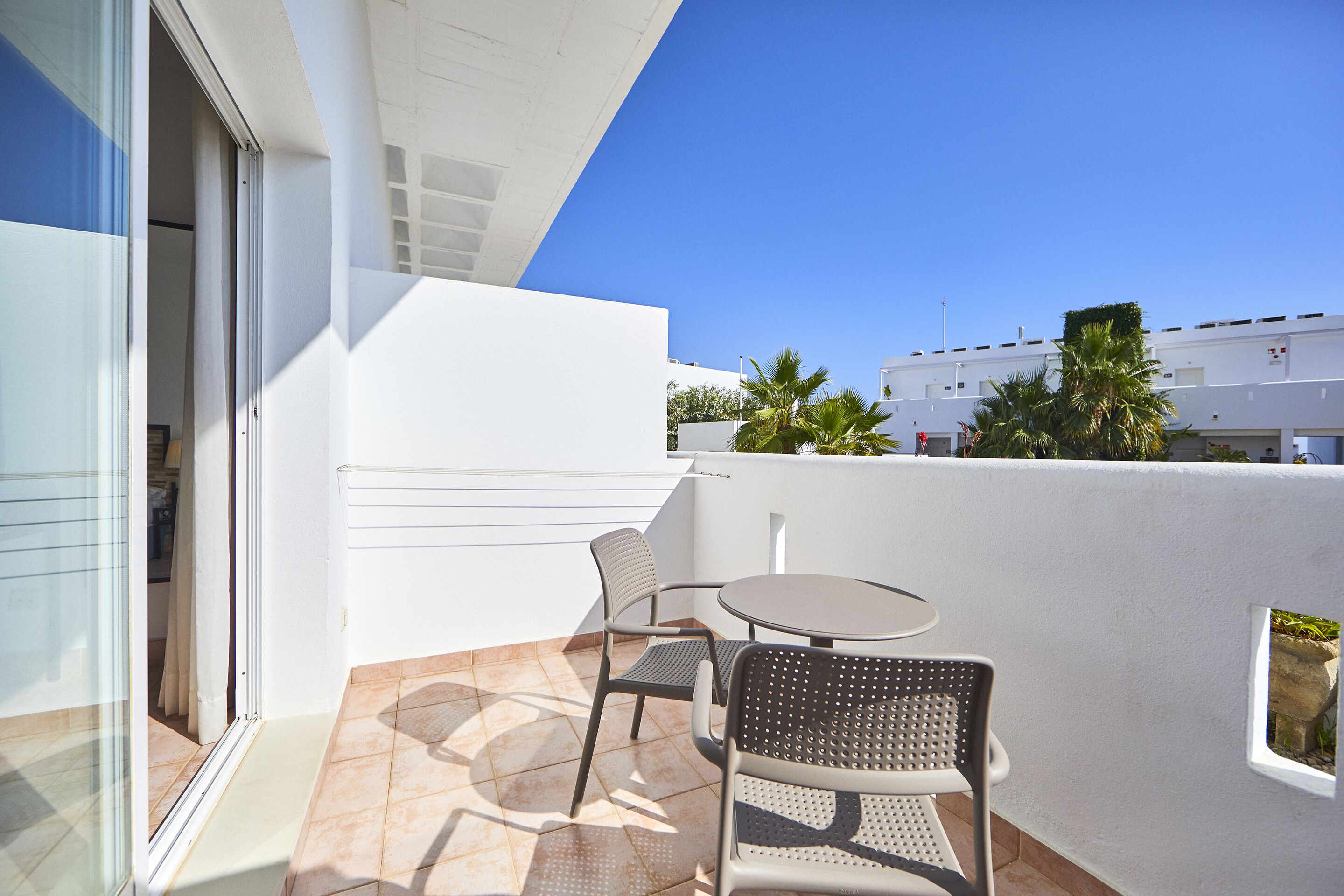 A balcony with a glass door, two chairs, a small round table, and a view of palm trees and buildings.