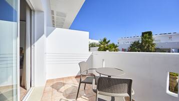 A balcony with a glass door, two chairs, a small round table, and a view of palm trees and buildings.