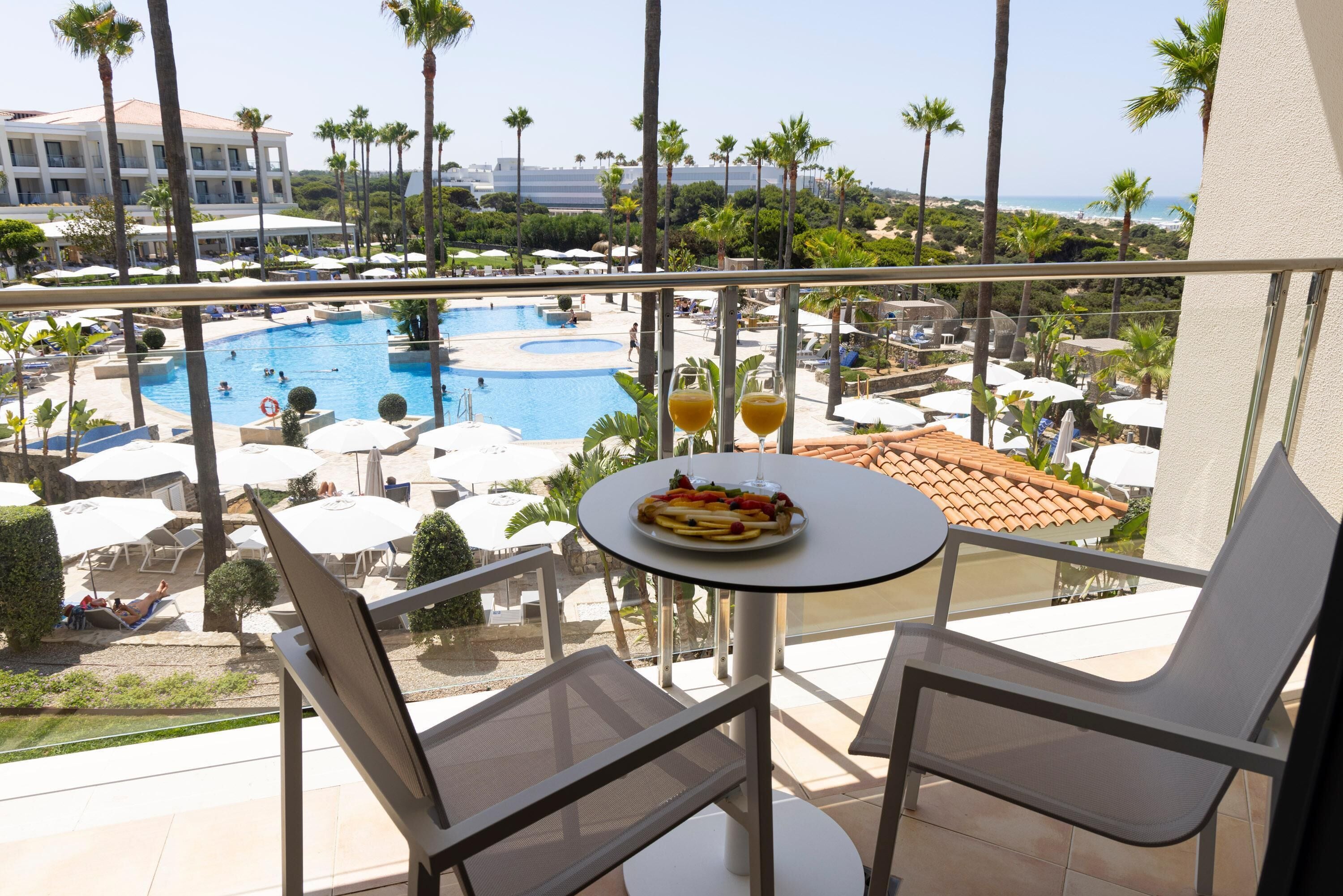 A balcony with a table and chairs overlooking a pool area with umbrellas and palm trees.