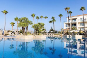A swimming pool with palm trees and lounge chairs, surrounded by a building with balconies. - Hipotels Barrosa Palace & SPA (Chiclana de la Frontera)
