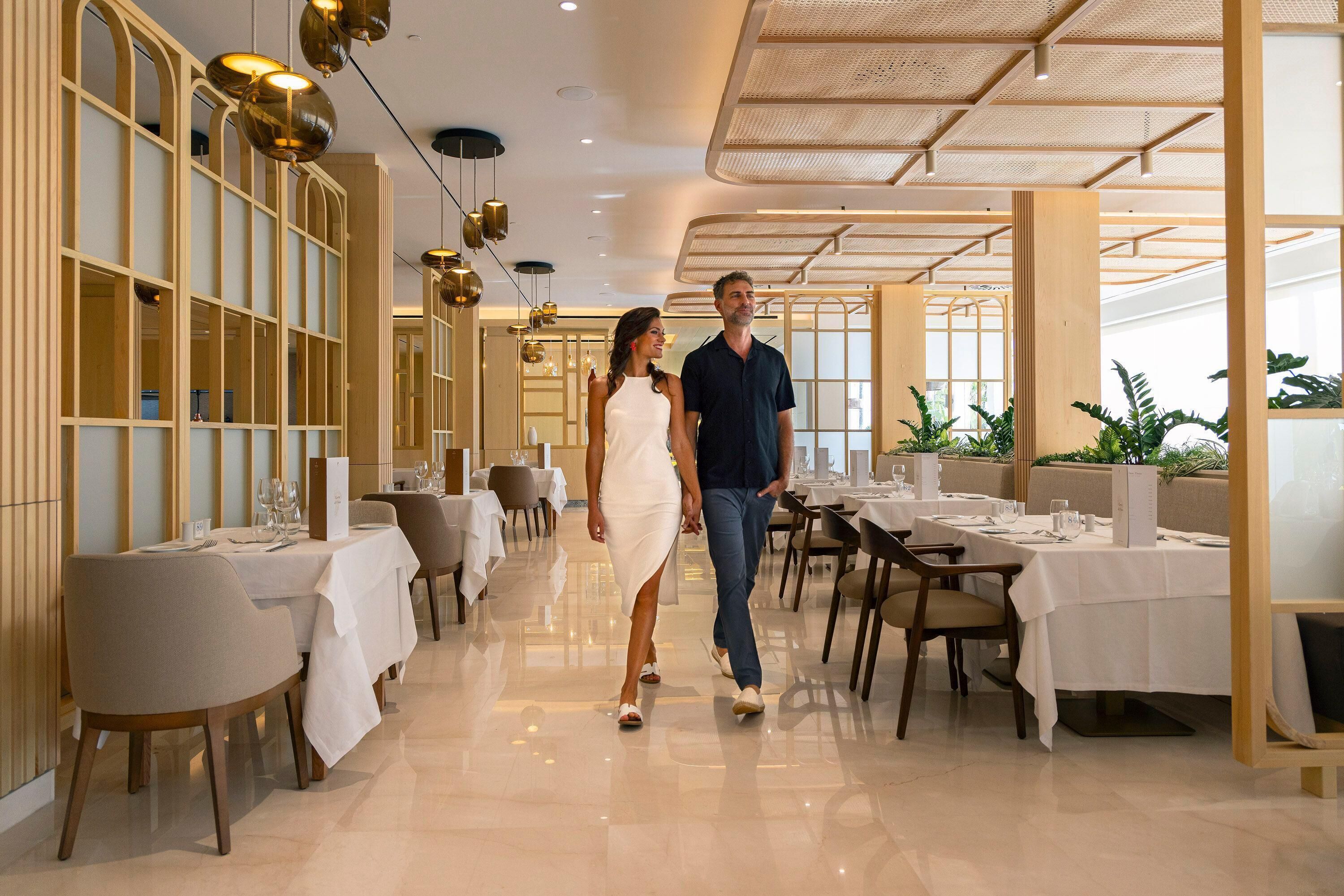 A spacious hotel corridor with tables and chairs, a high ceiling with hanging lights, and a modern design.