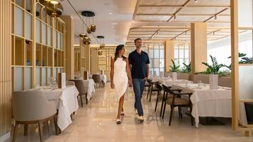 A spacious hotel corridor with tables and chairs, a high ceiling with hanging lights, and a modern design.