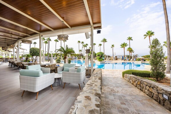 Un espacio al aire libre techado con mobiliario de mimbre, piscina, palmeras y paredes de piedra.