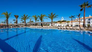 A large swimming pool with lounge chairs and palm trees, surrounded by hotel buildings.