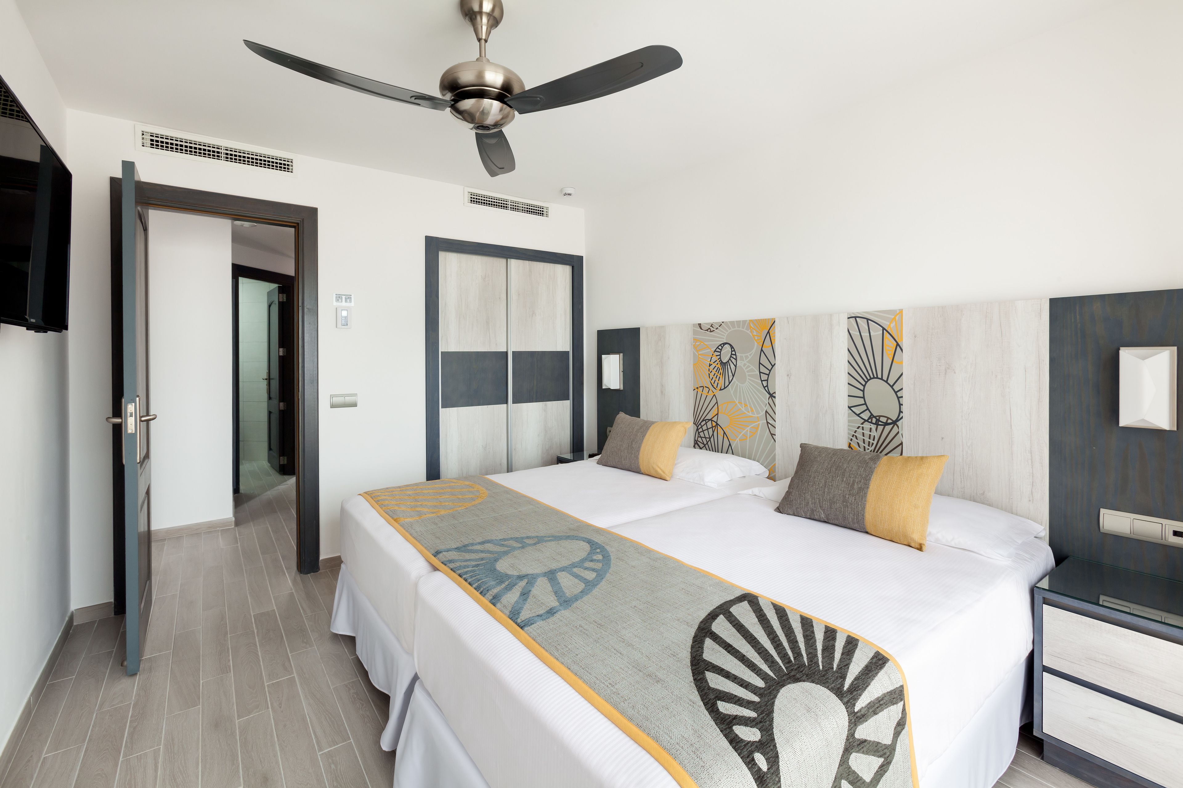 Habitación de hotel con una cama grande, ventilador de techo, mesita de noche y televisión instalada en la pared.