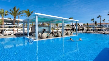 A swimming pool with lounge chairs and umbrellas, surrounded by palm trees and buildings.
