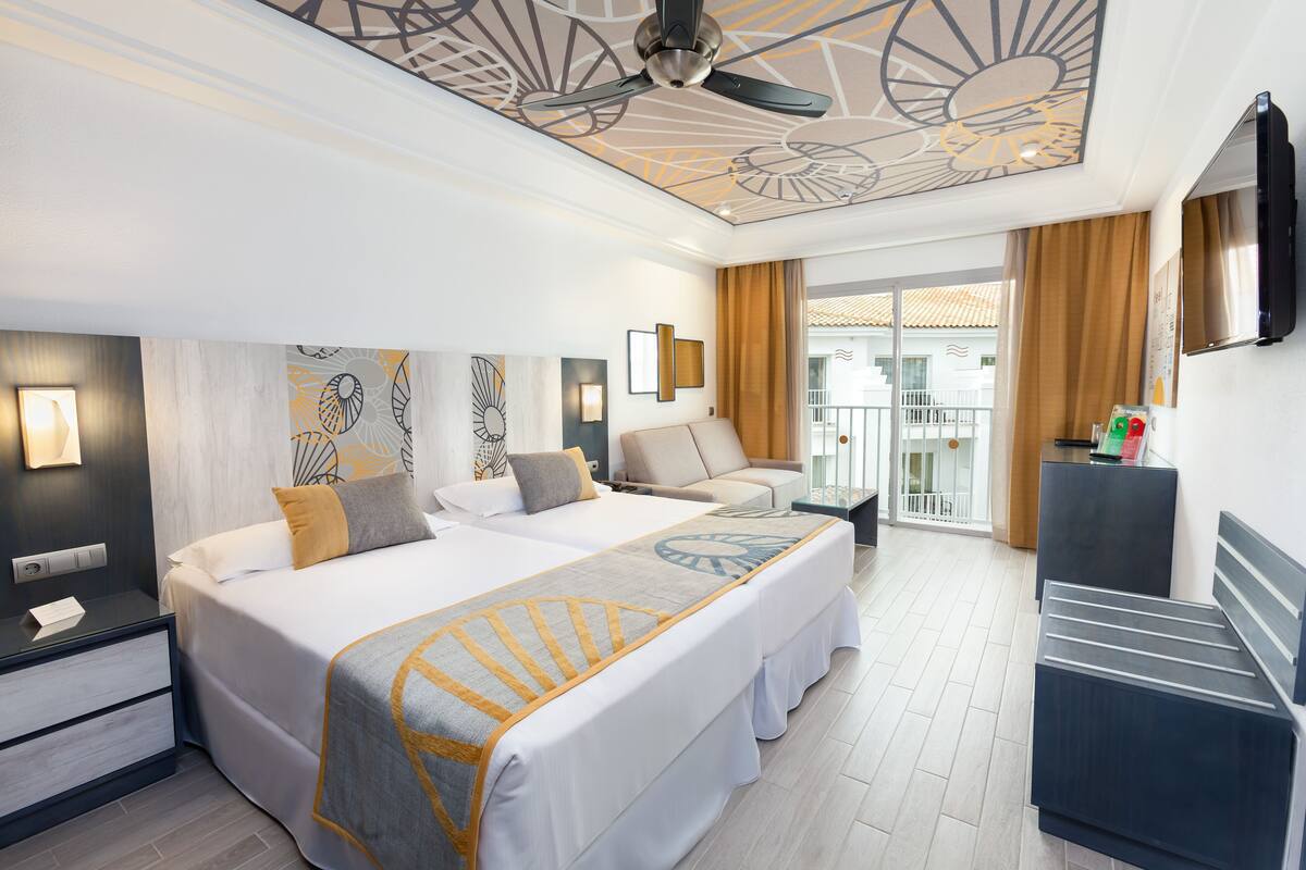 A hotel room with two beds, a ceiling fan, a TV, and a balcony with curtains.