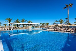 A clear blue pool with lounge chairs and umbrellas, surrounded by palm trees. - Hotel Riu Chiclana - All Inclusive (Chiclana de la Frontera)