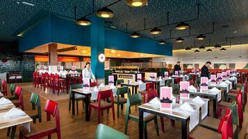 Un restaurante espacioso con varias mesas y sillas, un techo alto con luces colgantes y una zona de cocina al fondo.