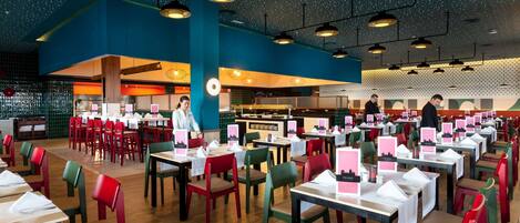 Un restaurante espacioso con varias mesas y sillas, un techo alto con luces colgantes y una zona de cocina al fondo.