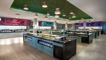 A modern buffet area with multiple serving stations, a central island with food displays, and a green ceiling with pendant lighting.
