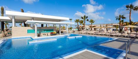 A swimming pool with lounge chairs and umbrellas, a bar area, and palm trees in the background.