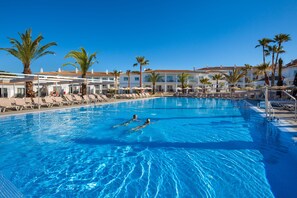 En swimmingpool med to personer, omgivet af liggestole og palmer, med hotelbygninger i baggrunden.