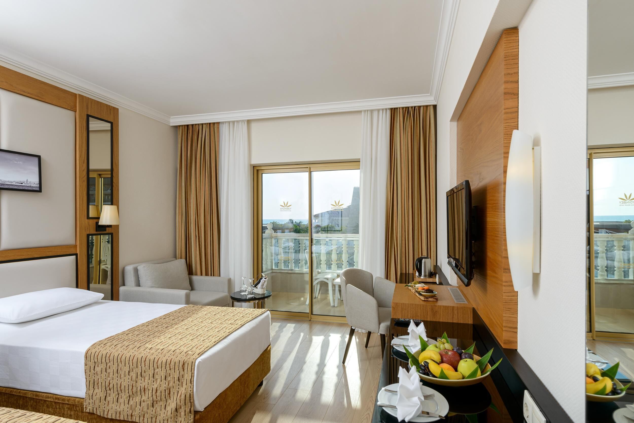 A hotel room with a bed, a desk with a TV, a balcony with a view, and a dining area with fruit.