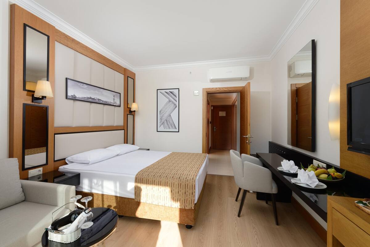A hotel room with a bed, a desk with a chair, a dining table with fruit, and a TV.