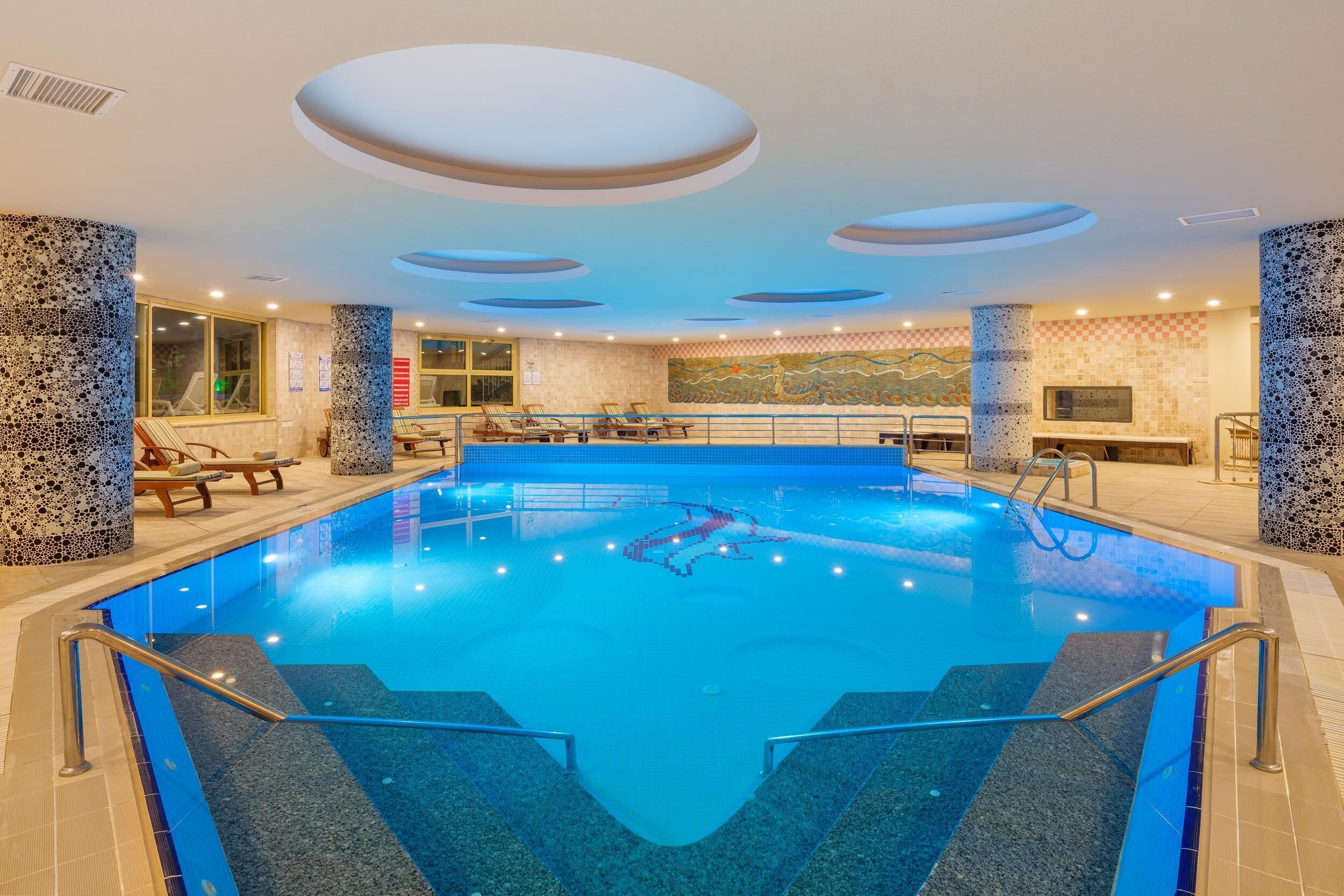 Una piscina interior con agua cristalina, rodeada de sillones de descanso y un suelo de baldosas.