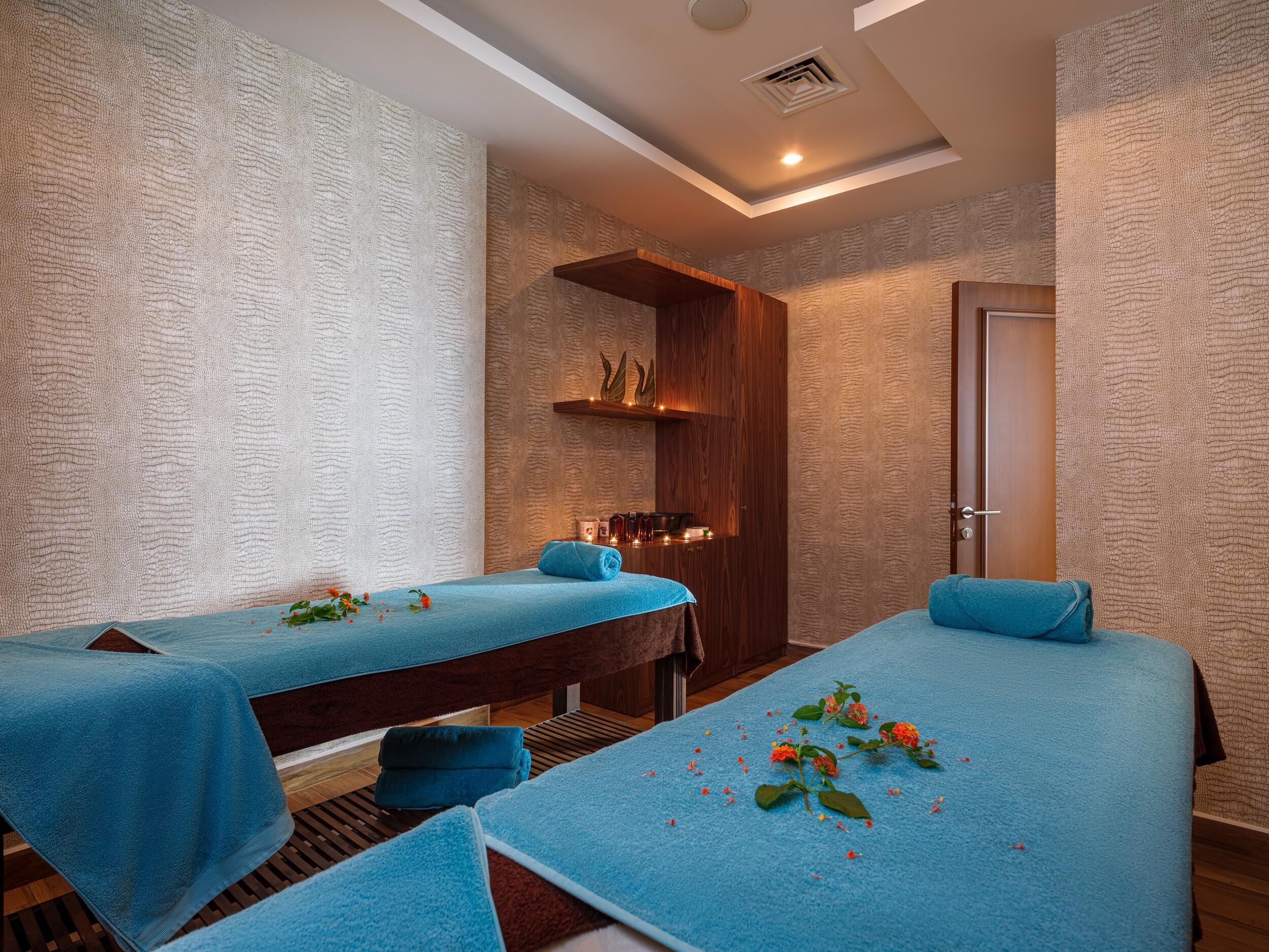A spa room with two massage tables, each covered with a blue towel. There are wooden benches and a shelf with candles and decorative items.