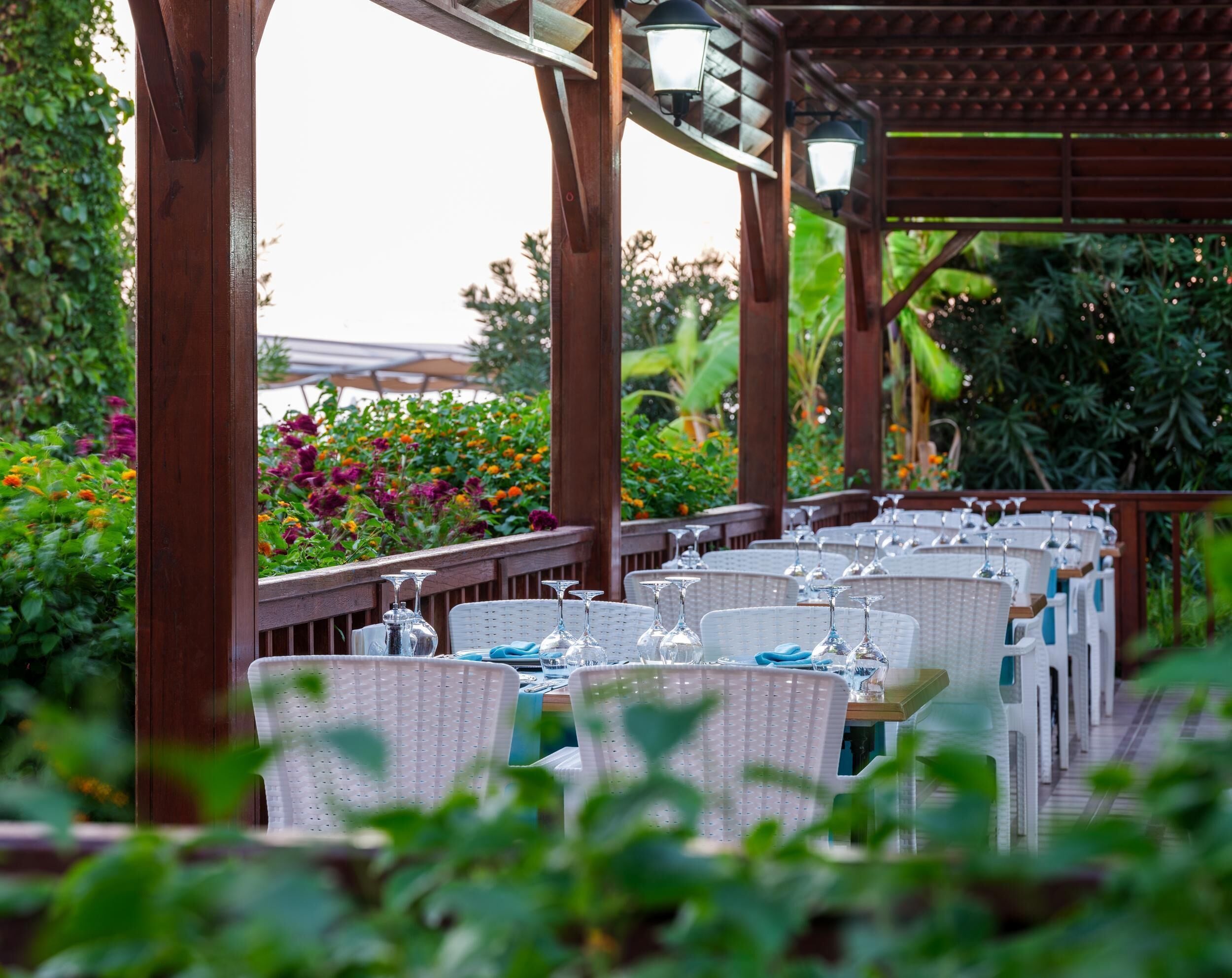 An outdoor dining area with wicker chairs and tables, surrounded by lush greenery and flowers.
