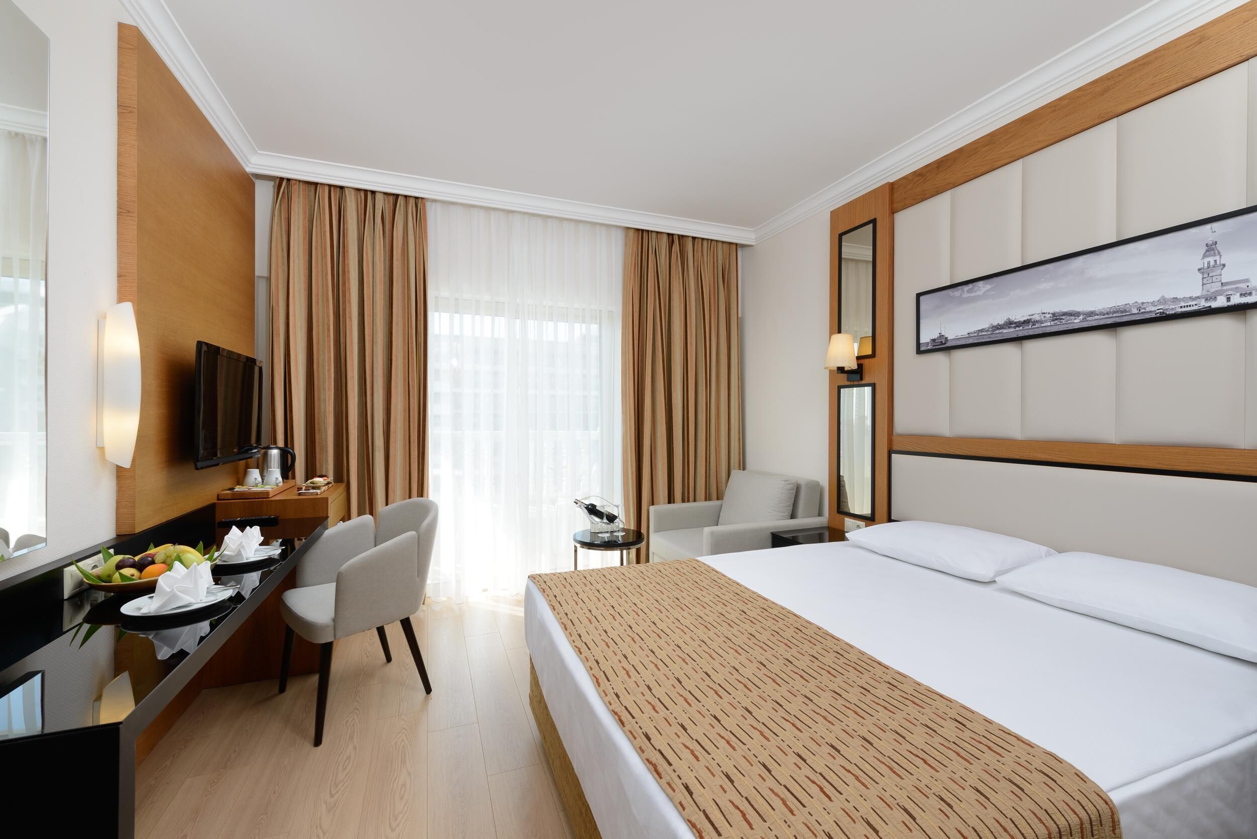 Habitación de hotel con cama, escritorio con computadora, silla, pequeña mesa con lámpara y ventana con cortinas.