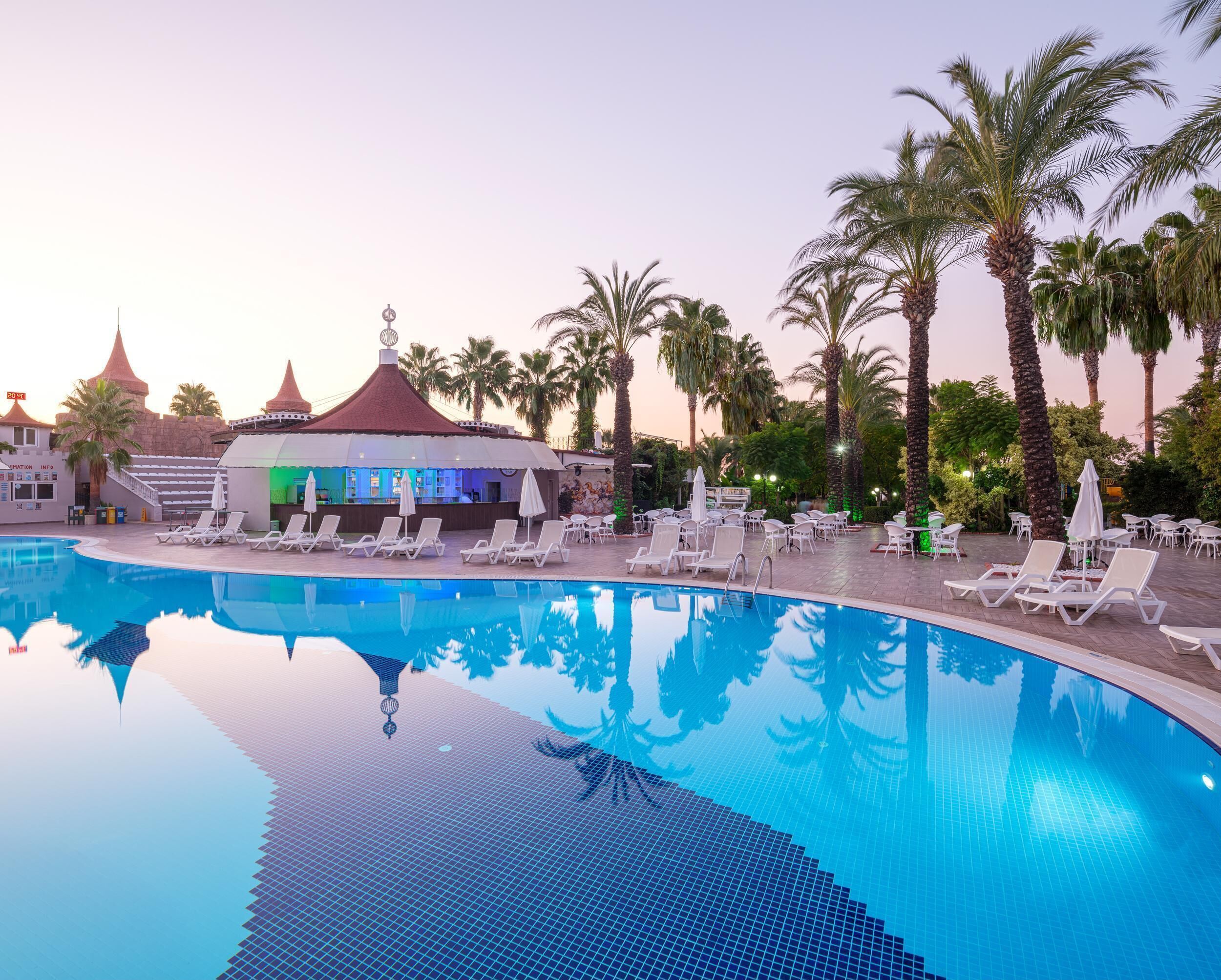 A swimming pool with lounge chairs and a bar area, surrounded by palm trees and a building with a red roof.
