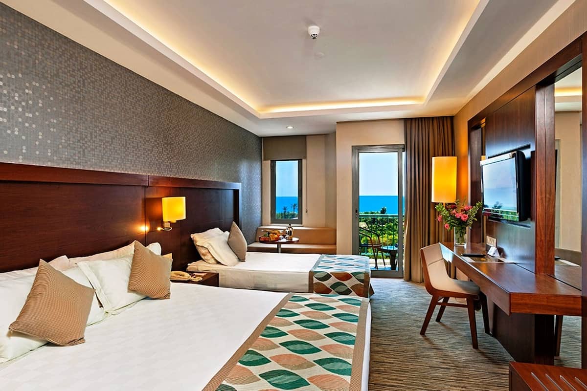 Ein Hotelzimmer mit zwei Betten, einem Schreibtisch, einem Fernseher und einem Balkon mit Meerblick.
