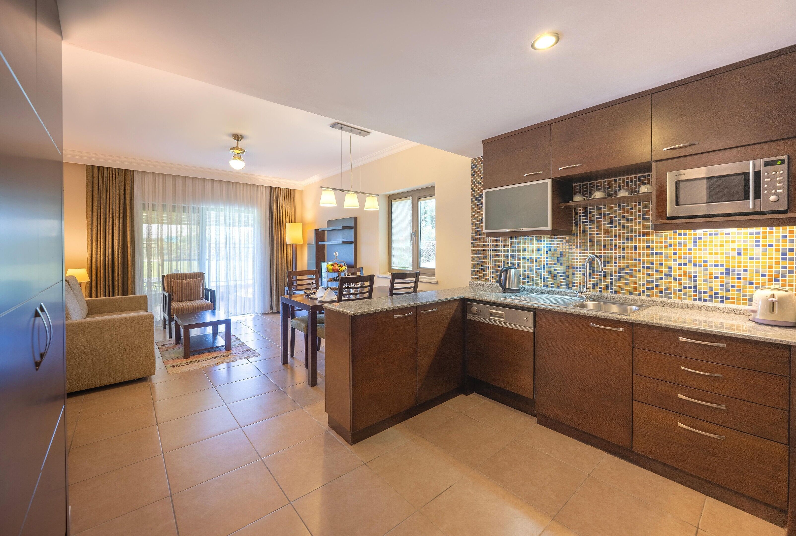 A modern kitchen with a microwave, stove, and sink, a dining area with chairs and a table, and a living area with a sofa and lamp.