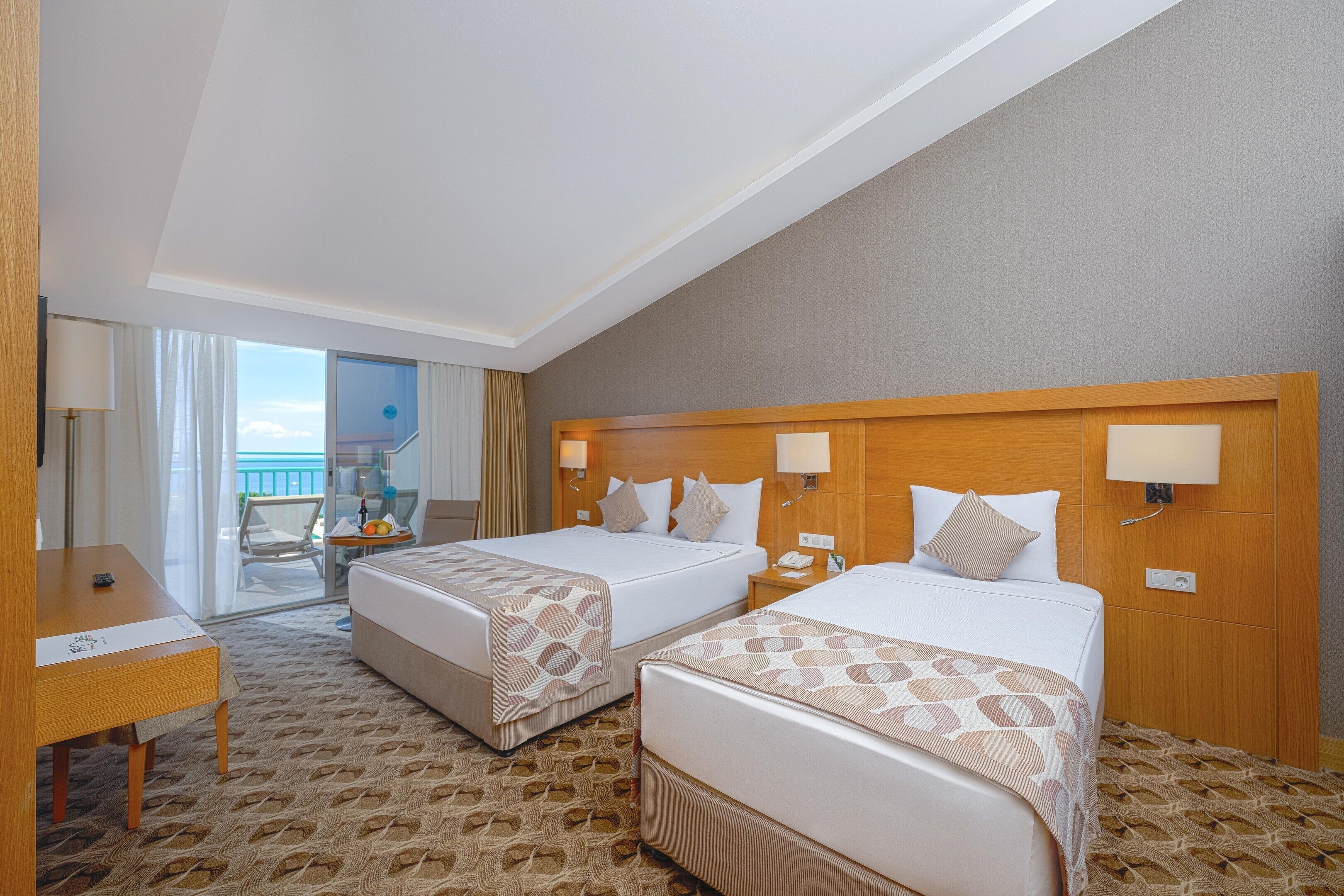 A hotel room with two beds, a desk, a chair, a balcony with a view, and a wooden headboard.
