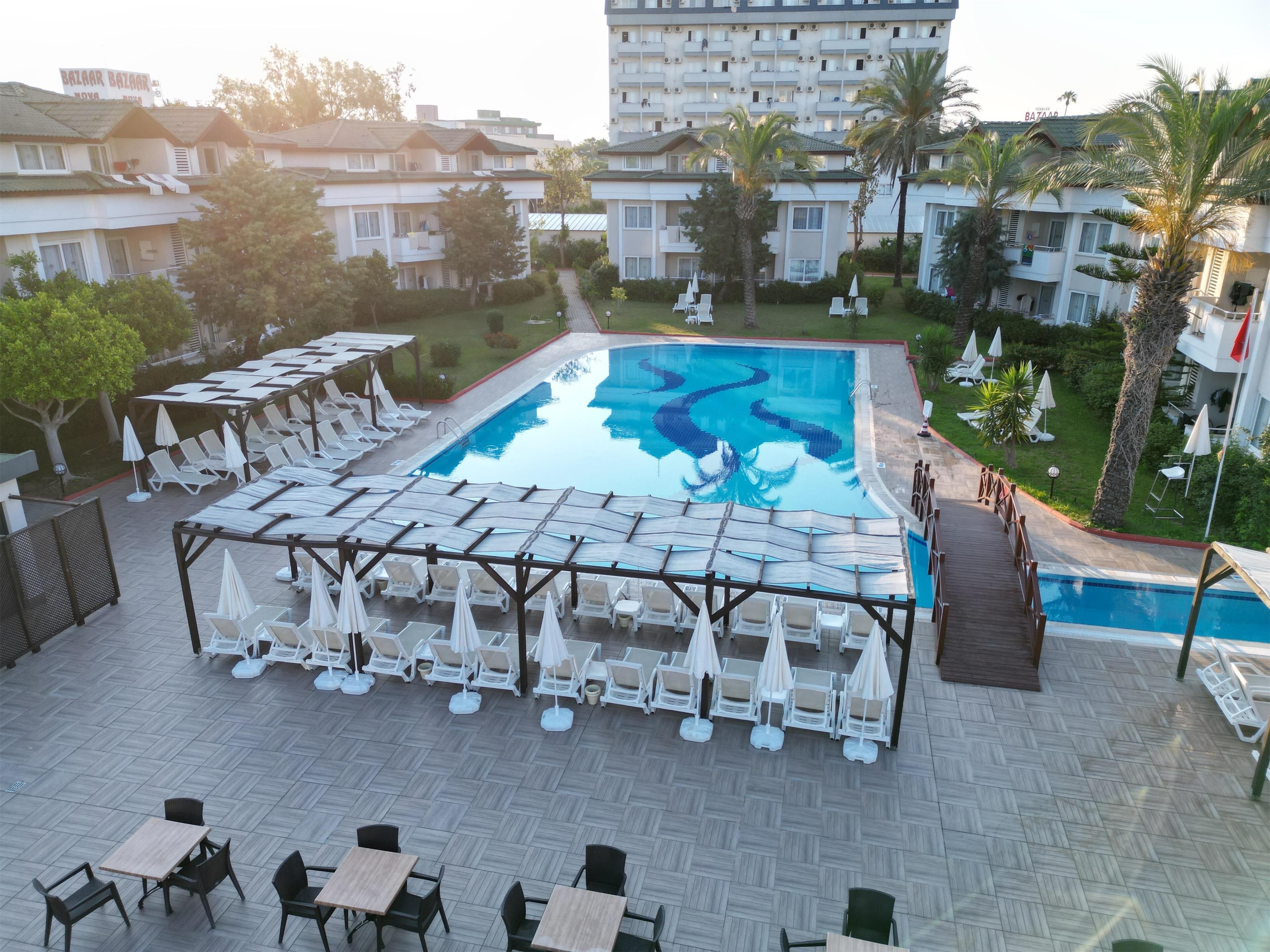 A pool area with lounge chairs and tables, surrounded by greenery and a multi-story building in the background.