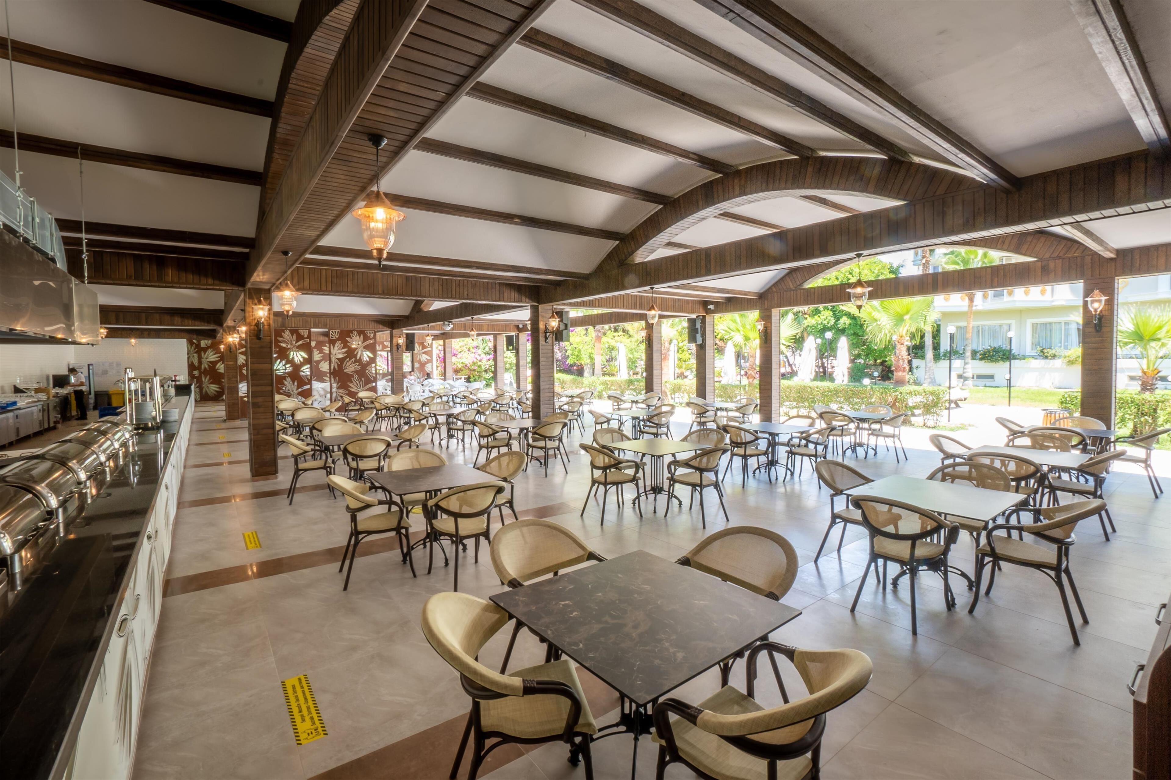 A spacious outdoor dining area with multiple tables and chairs, a covered section with a high ceiling, and a view of greenery outside.
