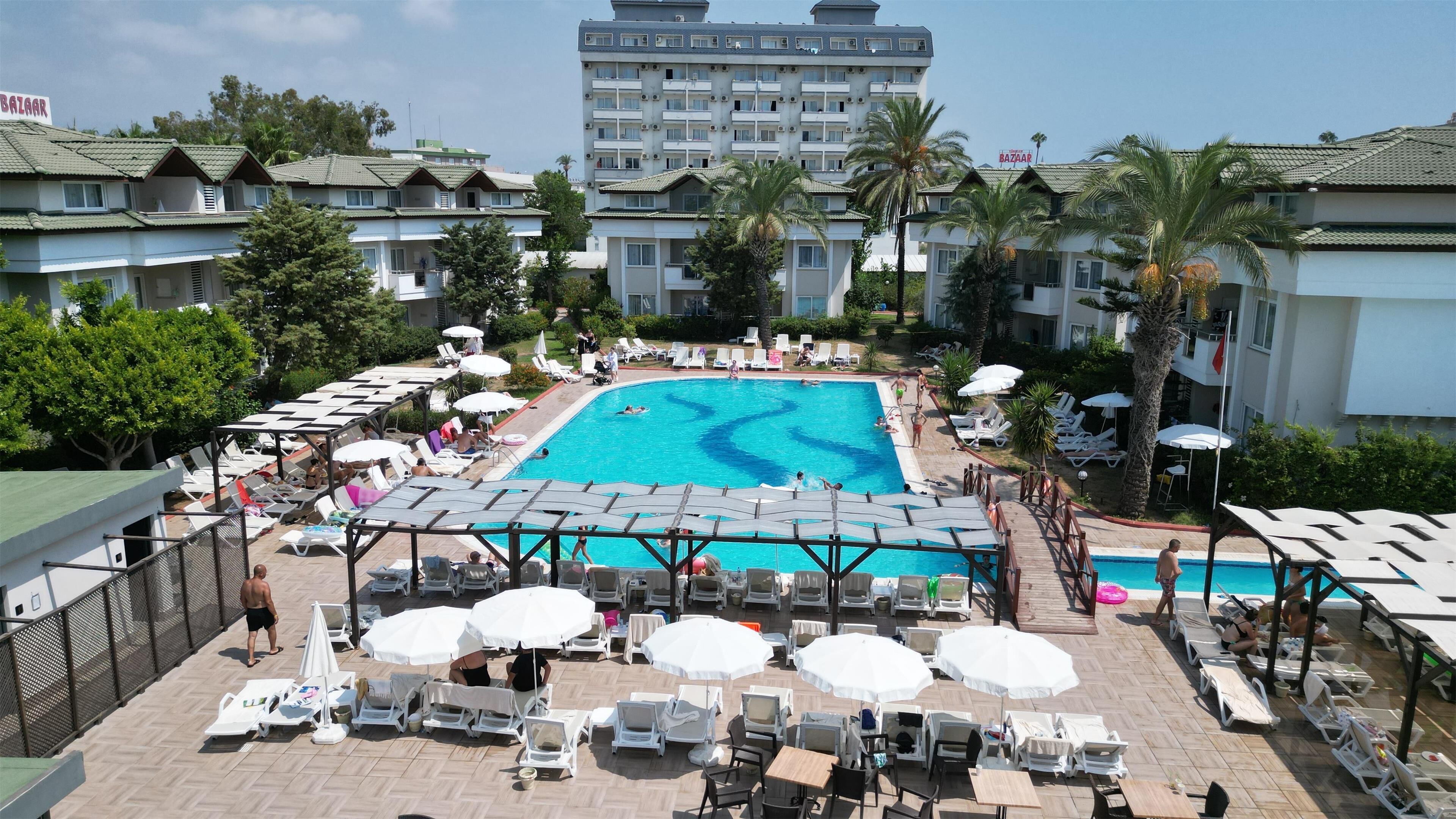 Zona de piscina de hotel con varias sillas de descanso y sombrillas, rodeada de palmeras y un edificio grande al fondo.